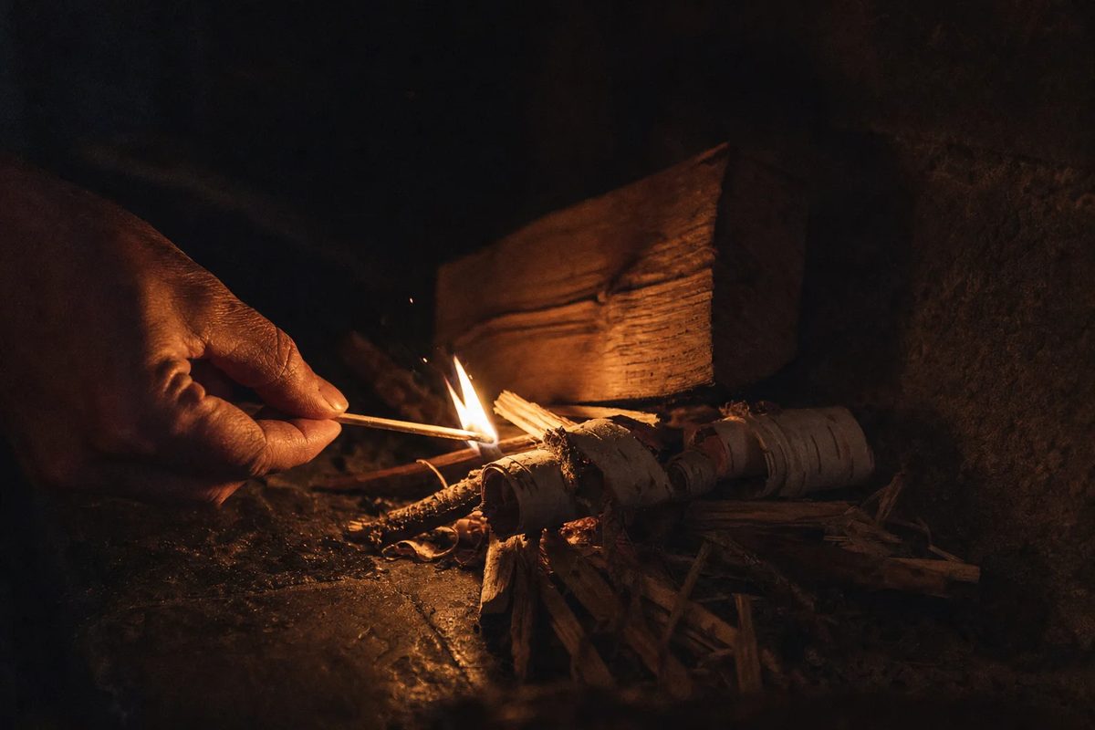 A man's hand holding a lit match to birch bark and dry kindling, a split log waiting behind it.