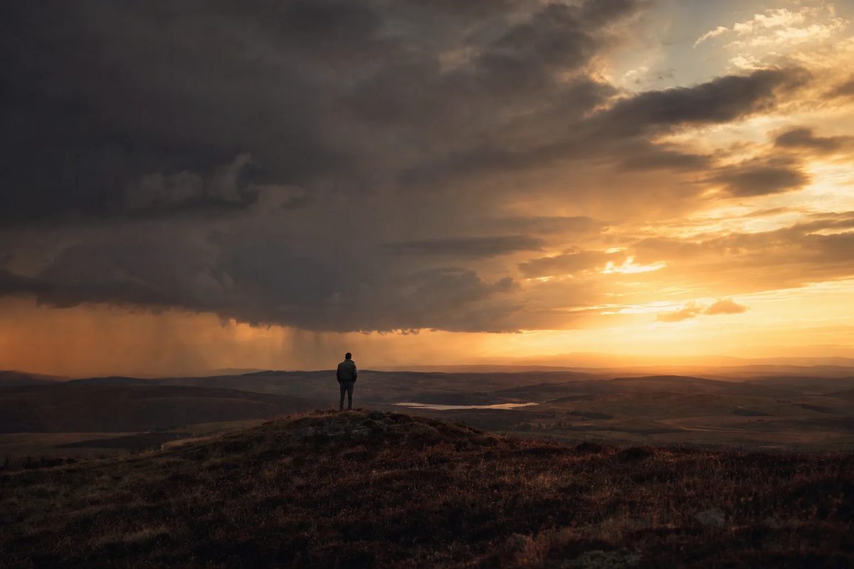 A vast dusk sky, storm cloud on one side and low gold light on the other, with one small figure standing on a rise of open moor.