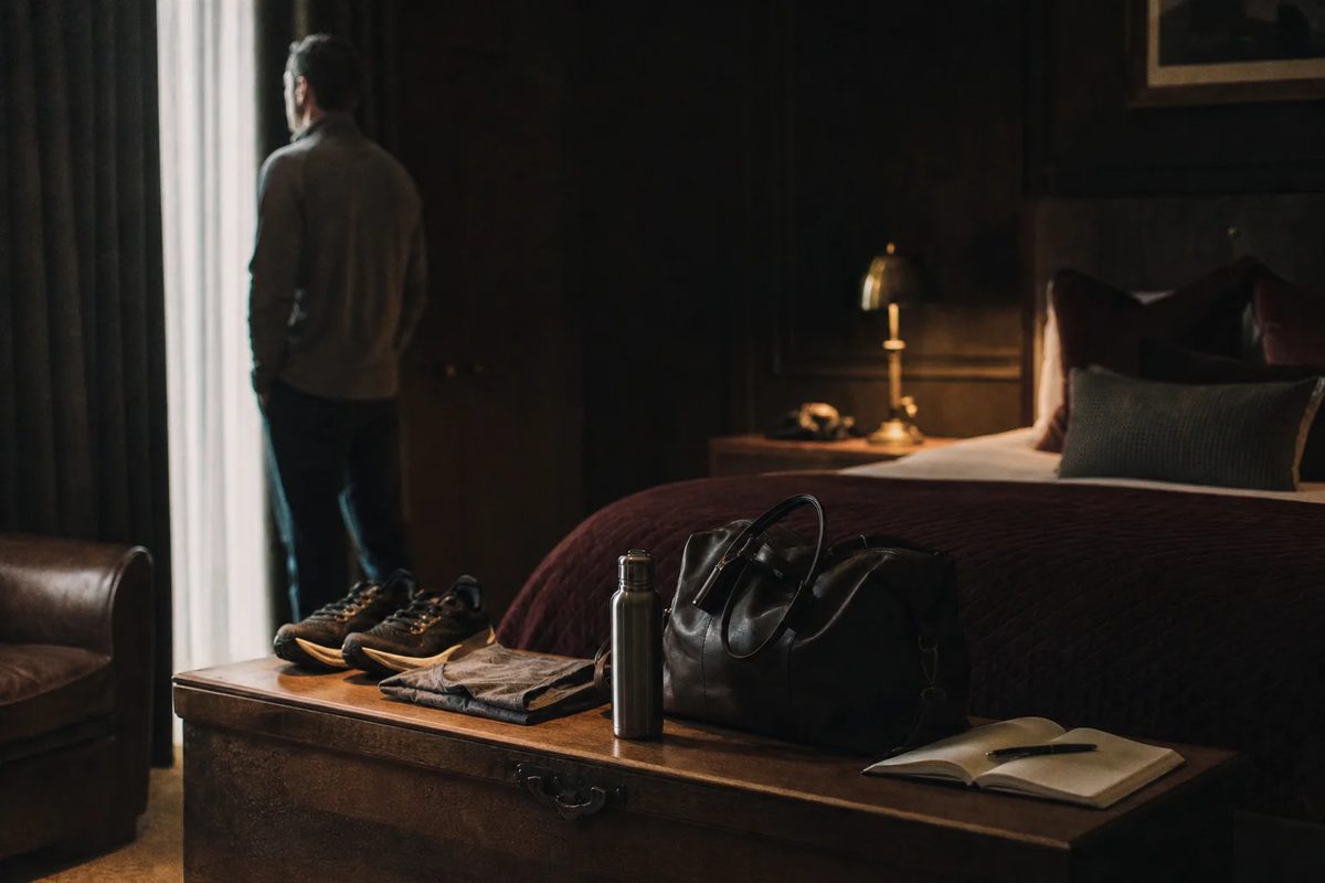 First light in a dark bedroom. Along the oak chest at the foot of the bed, a man's day is set out in a row: running shoes, a folded shirt, a flask, a work bag and an open notebook.
