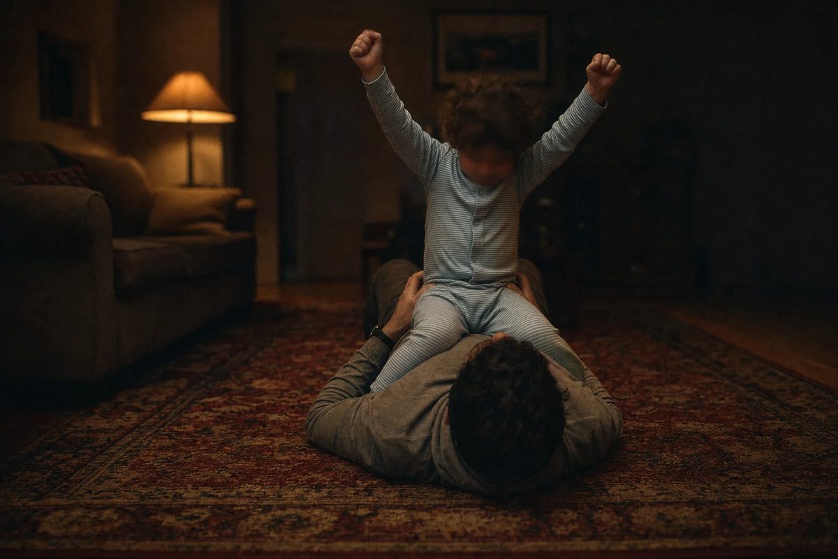 A lounge-room rug in lamplight, seen from above a man's shoulder as he lies flat on his back. A small child in soft focus sits on his chest with both arms raised.